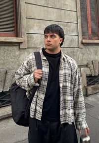 Jeune homme portant une chemise à carreaux et un t-shirt noir, avec un sac à dos noir, debout sur un trottoir en ville près d'un bâtiment en pierre.