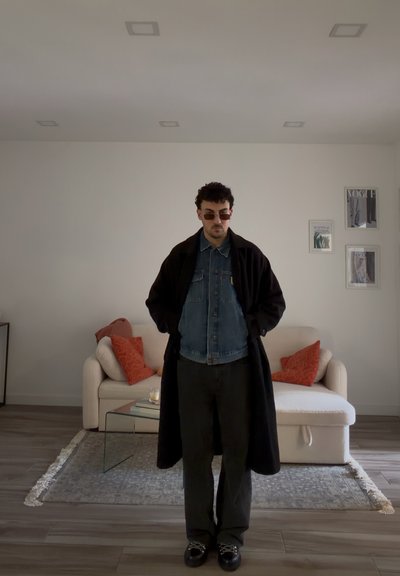 Hombre con gafas de sol, abrigo negro largo y chaqueta vaquera, de pie en una sala de estar con un sofá beige y cojines rojos.