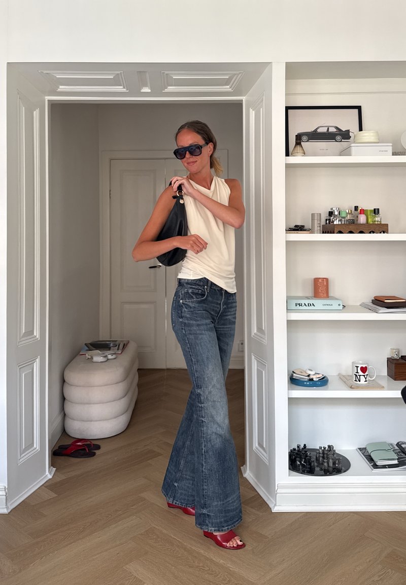 Femme portant des lunettes de soleil, un haut blanc sans manches, un jean évasé et des sandales rouges, debout dans l'embrasure d'une porte tenant un sac à main noir dans une pièce moderne.
