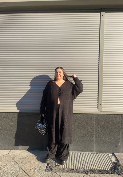 Cárdigan de punto negro con patrón de trenzas, abierto al frente y mangas largas. Combinado con pantalones negros de pierna ancha y un bolso de lunares.