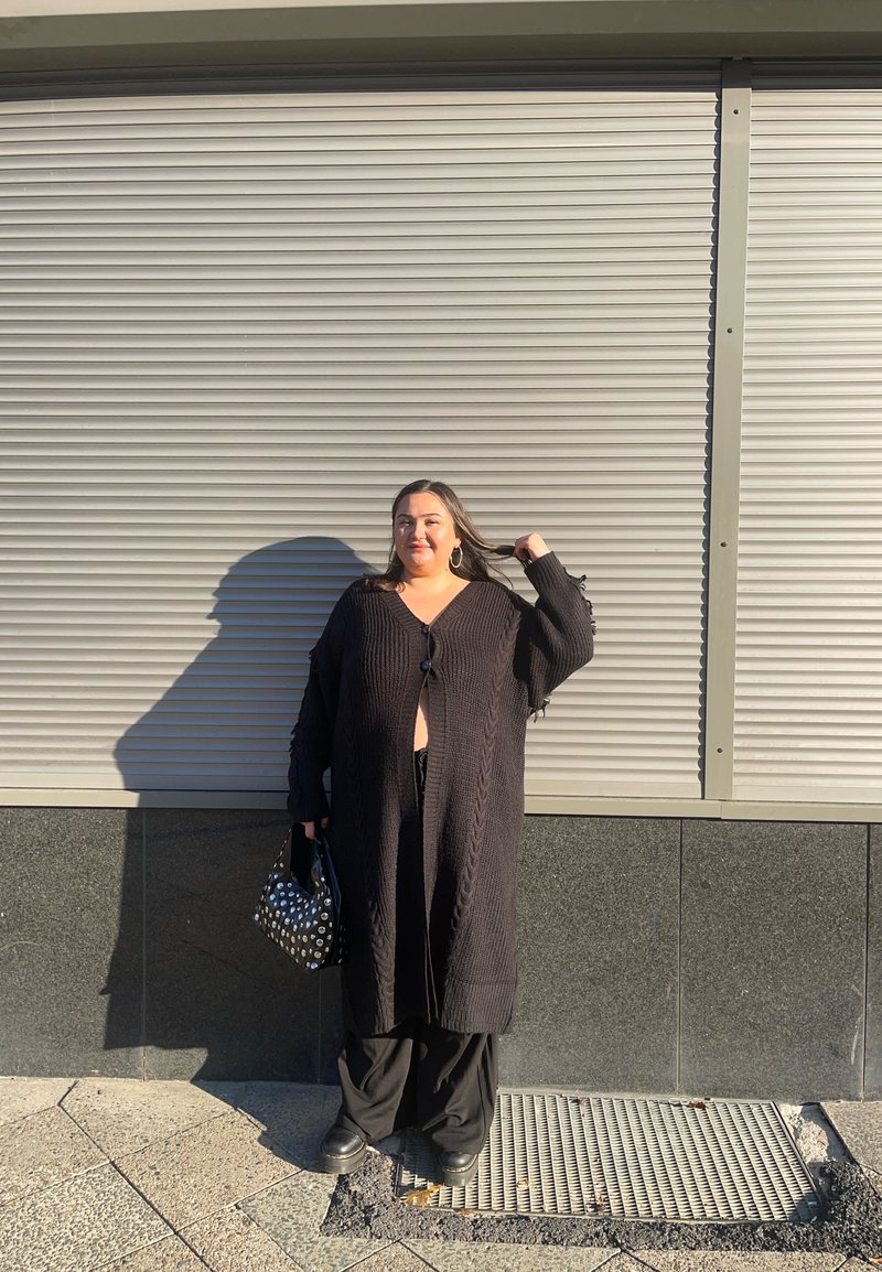 Black knit cardigan with a cable pattern, open front, and long sleeves. Paired with wide-leg black trousers and a polka dot bag.