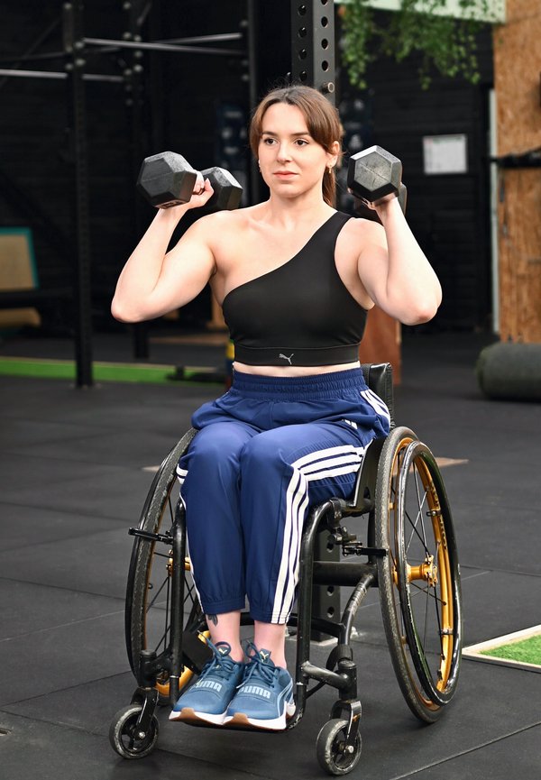 Γυναίκα σε αναπηρικό καροτσάκι σηκώνει αλτήρες μέσα σε κλειστό χώρο, φορά αθλητικά ρούχα, συγκεντρωμένη στην προπόνηση ενδυνάμωσης σε γυμναστήριο.