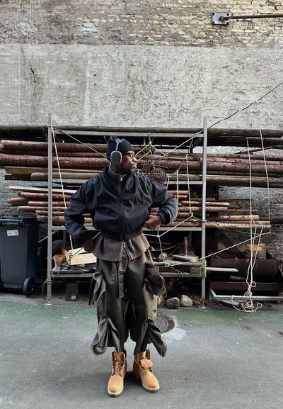 Chaqueta bomber negra con mangas anchas, suéter en capas y pantalones cargo verdes oliva con detalles de lazo exagerados; botas de trabajo amarillas completan el look.