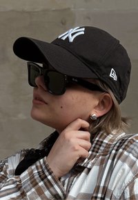 Czarna czapka z białym logo "NY", duże czarne okulary przeciwsłoneczne, srebrna kolczyk oraz brązowa koszula w kratkę z kontrastującą białą strukturą.