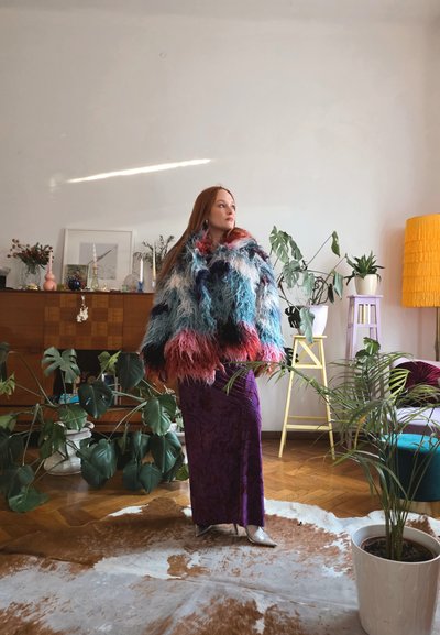 Mujer con cabello largo y rojo lleva una chaqueta peluda multicolor y falda morada, de pie en una habitación llena de plantas con suelo de madera y lámpara amarilla.