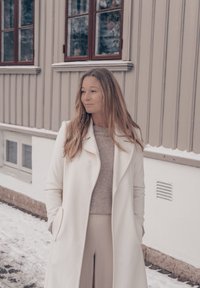 Femme aux longs cheveux portant un manteau blanc et une tenue beige, debout les mains dans les poches devant un bâtiment avec de la neige au sol.