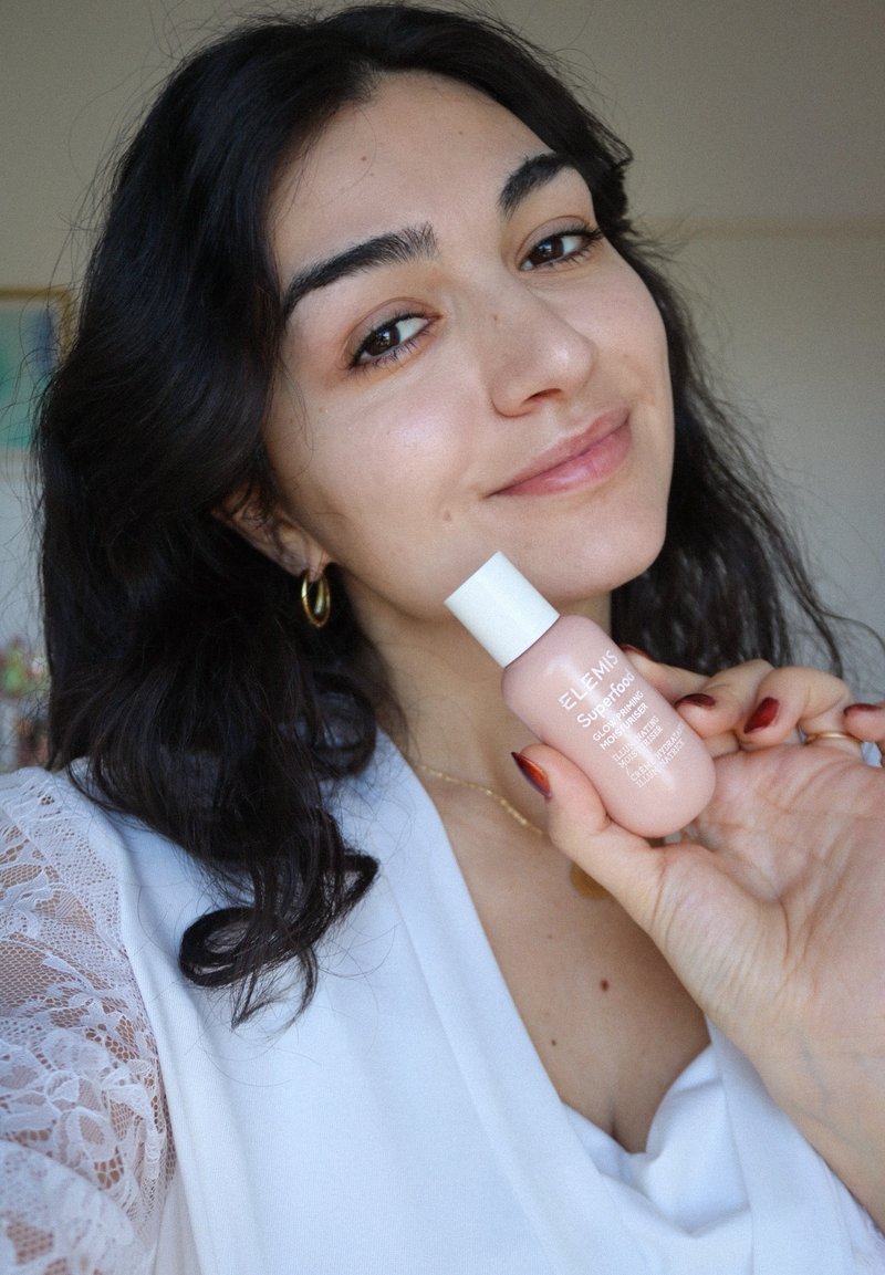 Roze flesje van ELEMIS Superfood Glow Priming Moisturizer in de hand gehouden. Plastic fles met een witte dop, gladde textuur, voorzien van een gelabeld ontwerp.