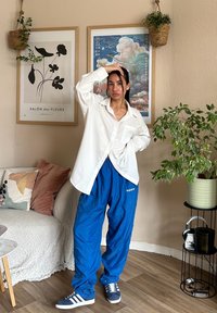 Λευκό πουκάμισο με μακριά μανίκια και κουμπιά, μπλε χαλαρές φόρμες με γράμματα και γκρι αθλητικά παπούτσια. Εσωτερικός χώρος με φυτά και έργα τέχνης.