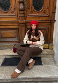 Pantalon en velours côtelé marron, pull en laine crème avec motif losanges, béret rouge, chaussons beige et sac à main bordeaux. Fond porte en bois.