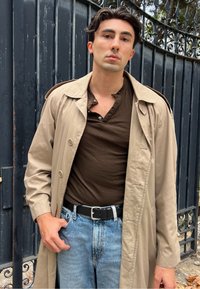 Beige színű trench coat, dupla gombolással, barna henley póló, világoskék farmernadrág és fekete öv egy textúrált vaskapun.