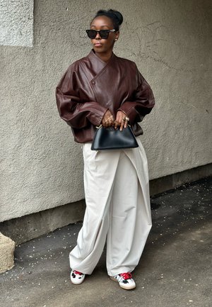 Mujer con chaqueta de cuero marrón de gran tamaño, pantalones anchos blancos, gafas de sol negras, sosteniendo un bolso negro, de pie contra una pared gris texturizada.