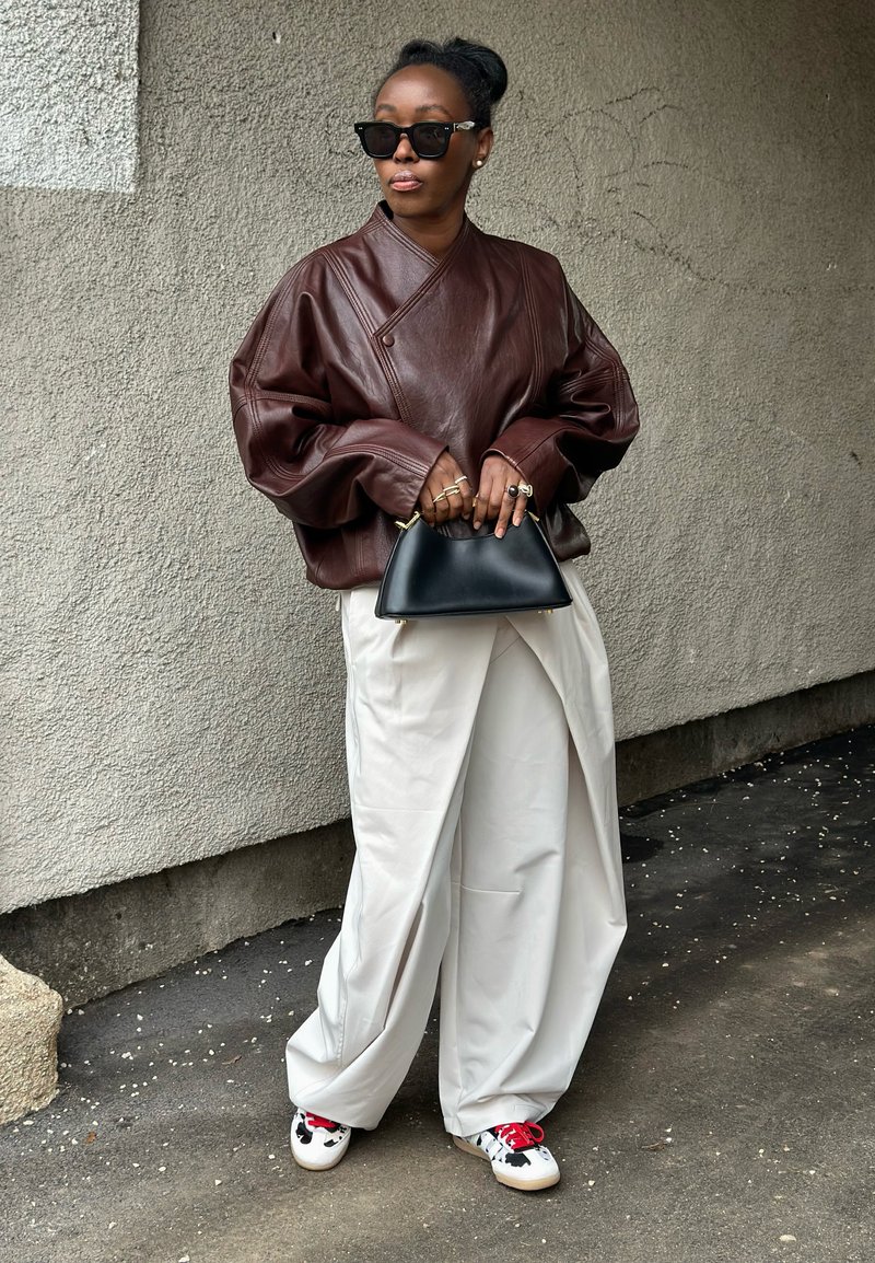 Femme portant une veste en cuir marron surdimensionnée, un pantalon blanc large, des lunettes de soleil noires, tenant un sac à main noir, debout devant un mur gris texturé.