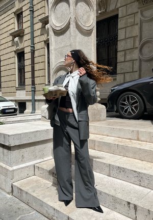Blazer gris a rayas y pantalones de pierna ancha a juego con un top corto negro. Sosteniendo un envase de comida, con una diadema negra y pendientes.