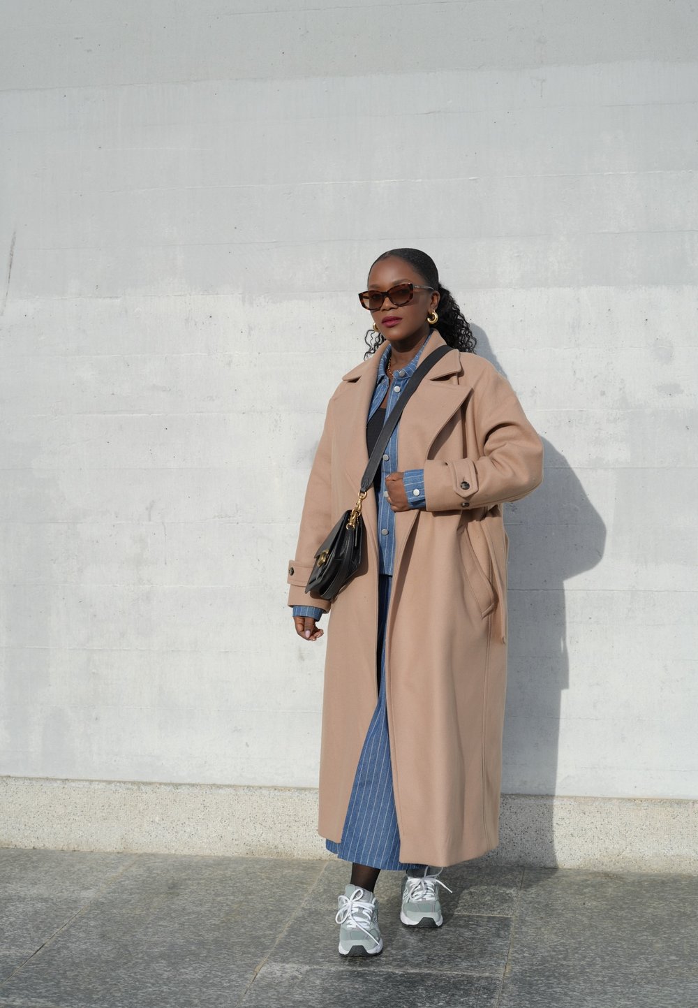 Μπεζ μακρύ παλτό, τζιν πουκάμισο από κάτω, φούστα με κατακόρυφες ρίγες, γκρι και λευκά αθλητικά παπούτσια, μαύρη διαγώνια τσάντα, μεγάλα γυαλιά ηλίου.