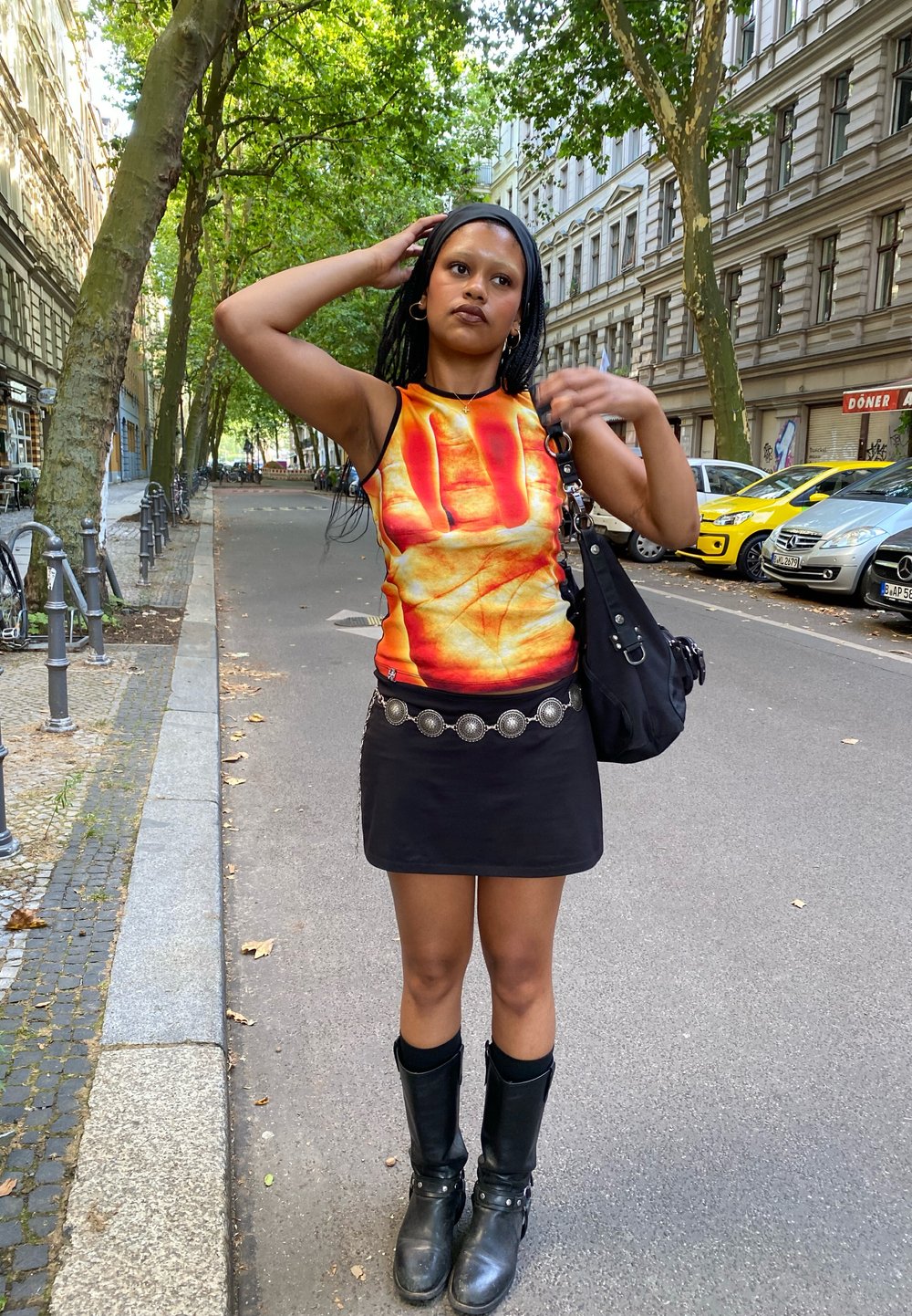 Top sem mangas com uma gráfica vibrante em vermelho e laranja, combinado com uma saia e cinto pretos. Botas pretas até ao joelho completam o visual.