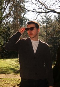 Jeune homme en plein air sous la lumière du soleil ajuste ses lunettes de soleil noires, portant un cardigan sombre par-dessus une chemise blanche, avec des arbres sans feuilles et une zone herbeuse en arrière-plan.