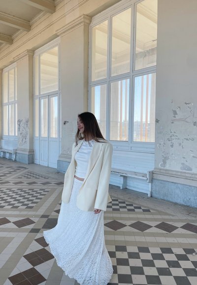 Maxifalda blanca de encaje con un patrón texturizado, combinada con una blusa blanca ajustada y un blazer color crema. El fondo presenta grandes ventanales y un suelo de azulejos.