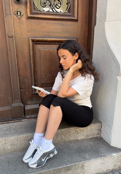 Una persona sentada con una camiseta blanca y leggings negros, sosteniendo un teléfono inteligente, con zapatillas plateadas y blancas, en escalones de piedra junto a una puerta de madera.