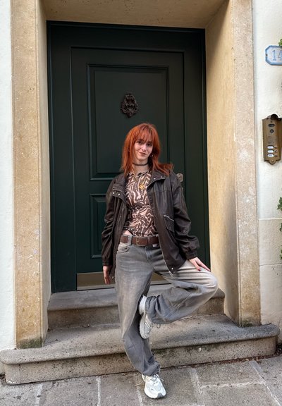 Chaqueta de cuero marrón sobre una blusa con estampado, pantalones gris holgados y zapatillas blancas. De pie sobre una pierna en unos escalones de piedra junto a una puerta verde.