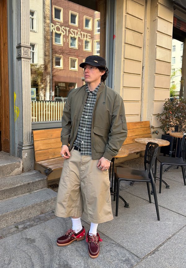 Hombre con sombrero negro, chaqueta verde, camisa de cuadros, pantalones cortos anchos beige, calcetines blancos y zapatos rojos, de pie fuera de una cafetería con bancos de madera.