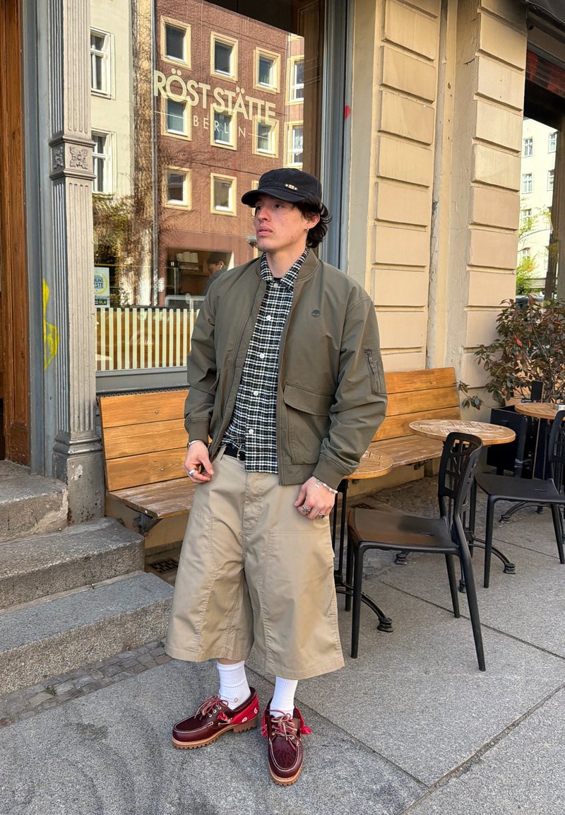 Homme portant un chapeau noir, une veste verte, une chemise à carreaux, un short large beige, des chaussettes blanches et des chaussures rouges, debout devant un café avec des bancs en bois.