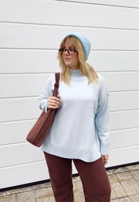 Femme aux cheveux blonds portant des lunettes, un pull bleu clair et un bonnet, un pantalon marron, et portant un sac à bandoulière marron, debout contre un mur blanc.
