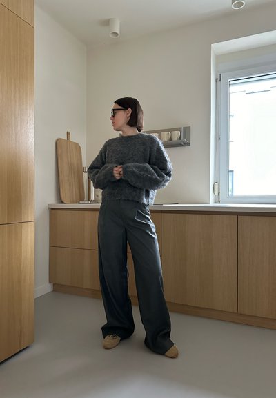 Suéter gris difuso con hombros caídos, combinado con pantalones sueltos gris oscuro y zapatos marrón claro, ubicado en una cocina moderna.