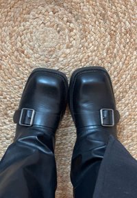 Sabots en cuir noir avec un bout carré et un détail de boucle sur le côté. Texture lisse, portés avec un pantalon noir ajusté, sur un fond tissé.