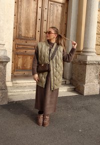 Chaleco acolchado verde oliva, vestido de lana marrón y botas marrón con cordones. Los accesorios incluyen un bolso cruzado marrón y gafas de sol.