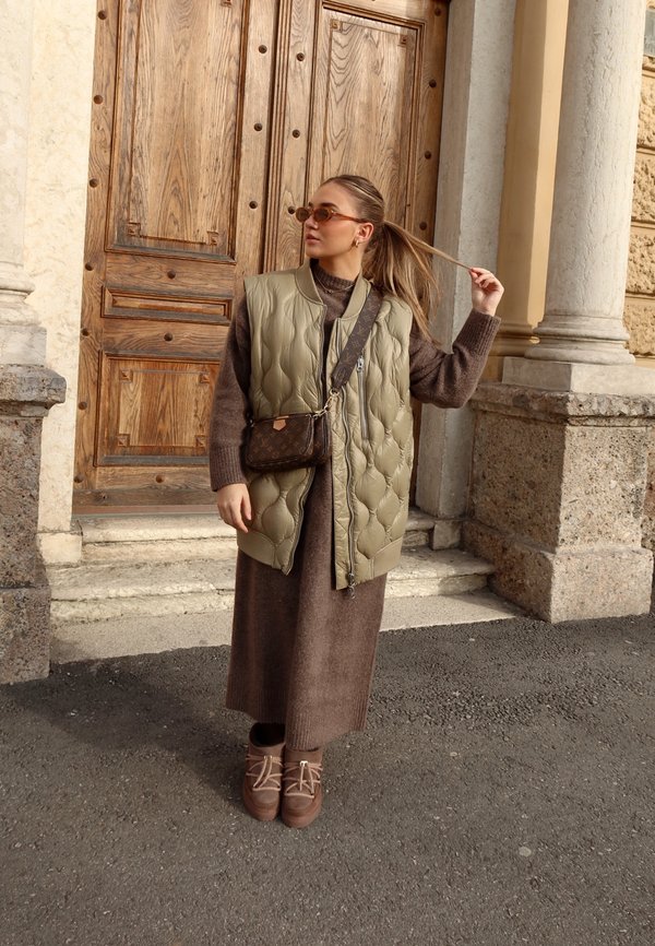 Chaleco acolchado verde oliva, vestido de lana marrón y botas marrón con cordones. Los accesorios incluyen un bolso cruzado marrón y gafas de sol.