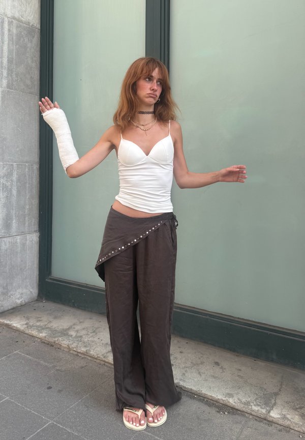Junge Frau mit schulterlangem Haar, die ein weißes Top und dunkle Hose trägt, steht mit einem Gips am linken Arm an einer Gebäudewand.