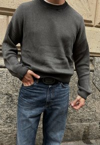 Man som bär en mörkgrå ribbad tröja och blå jeans med handen i fickan, står mot en stenvägg.