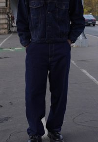 Mörk denimjacka och byxor med en avslappnad passform. Jackan har framfickor och knäppning med knappar. Båda plaggen har en enhetlig blå färg.