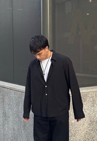 Schwarzes Hemd mit normaler Passform, getragen über einem weißen T-Shirt. Das Hemd hat einen Kragen und lange Ärmel und zeichnet sich durch eine glatte Textur aus.