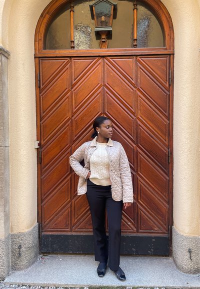 Chaqueta acolchada beige con cremallera, suéter de punto con trenzas crema, pantalones negros y zapatos negros, posando frente a una puerta de madera con patrón de espiga.