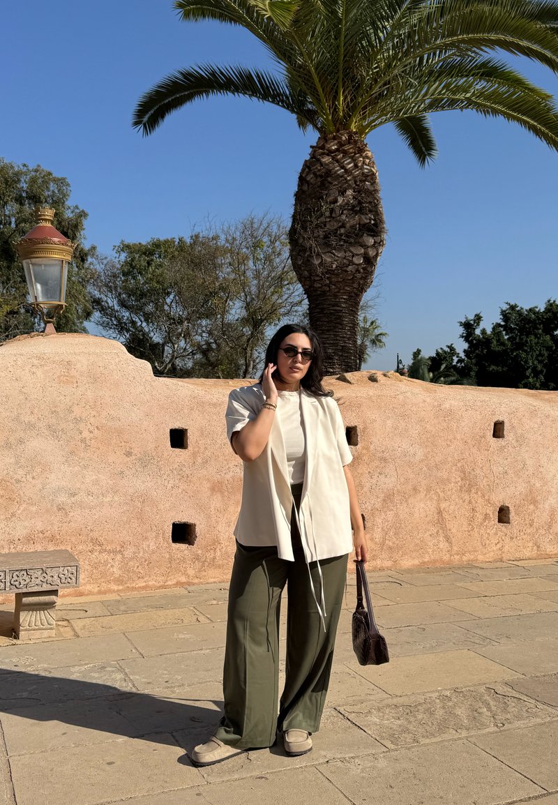 Donna con occhiali da sole, giacca color crema e pantaloni oliva in piedi su un pavimento in pietra accanto a un muro color sabbia con una palma e un lampione in un ambiente esterno soleggiato.