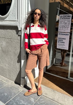 Mujer con cabello rizado, gafas de sol, suéter a rayas rojas y blancas, pantalones cortos marrones y zapatos marrones, está de pie fuera de una tienda bajo la luz solar.