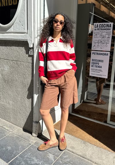 Mujer con cabello rizado, gafas de sol, suéter a rayas rojas y blancas, pantalones cortos marrones y zapatos marrones, está de pie fuera de una tienda bajo la luz solar.