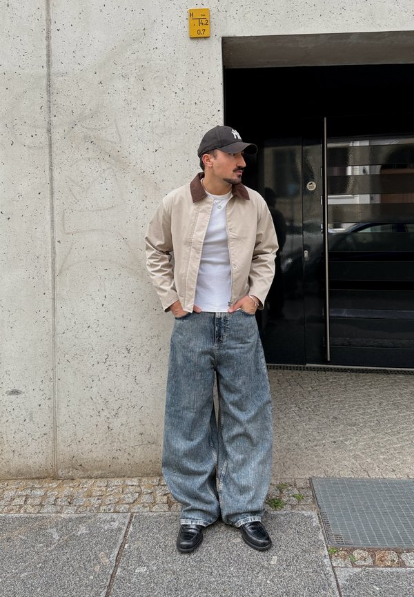 Homme portant un jean large bleu, une veste beige avec un col marron, un t-shirt blanc, une casquette noire et des chaussures noires, debout près d'un mur en béton et d'une porte sombre.