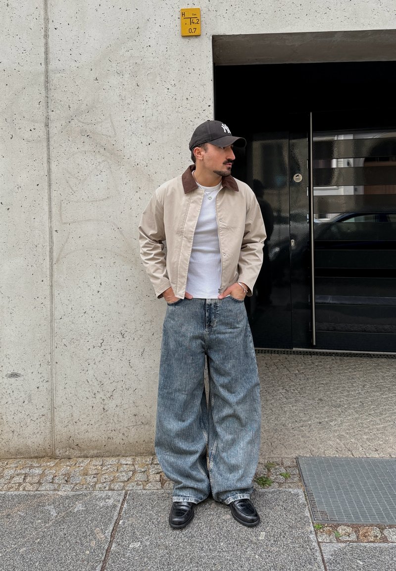 Homme portant un jean large bleu, une veste beige avec col marron, un t-shirt blanc, une casquette noire et des chaussures noires, debout près d'un mur en béton et d'une porte sombre.