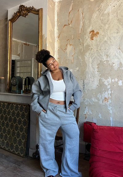 Joven mujer en pantalones de chándal grises y sudadera se reclina con los ojos cerrados en una habitación rústica con paredes desconchadas y un gran espejo ornamental.