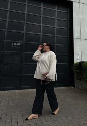 Suéter beige de gran tamaño, pantalones de pierna ancha negros, bolso marrón y zapatos planos bicolor. De pie frente a una puerta de garaje con un patrón de rejilla negra.