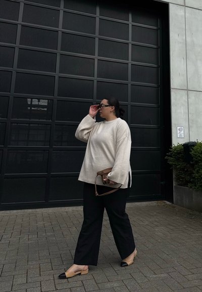 Suéter beige de gran tamaño, pantalones de pierna ancha negros, bolso marrón y zapatos planos bicolor. De pie frente a una puerta de garaje con un patrón de rejilla negra.