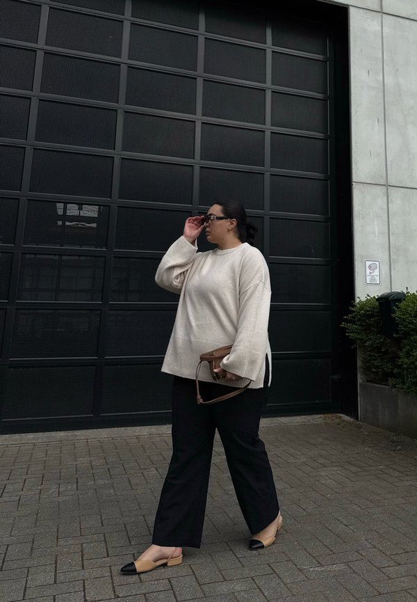 Beige oversized sweater, sorte brede bukser, brun håndtaske og tofarvede flade sko. Står foran en sort gittermønster garageport.