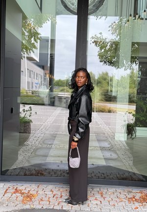 Chaqueta de cuero negra, pantalones largos negros, pulsera con detalles plateados, sosteniendo un pequeño bolso brillante. Puerta de cristal y pasarela pavimentada en el fondo.