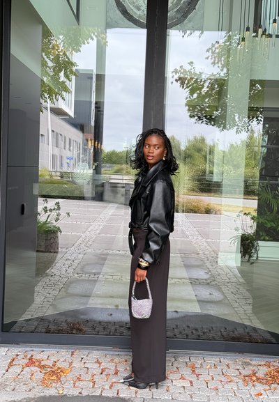 Chaqueta de cuero negra, pantalones largos negros, pulsera con detalles plateados, sosteniendo un pequeño bolso brillante. Puerta de cristal y pasarela pavimentada en el fondo.