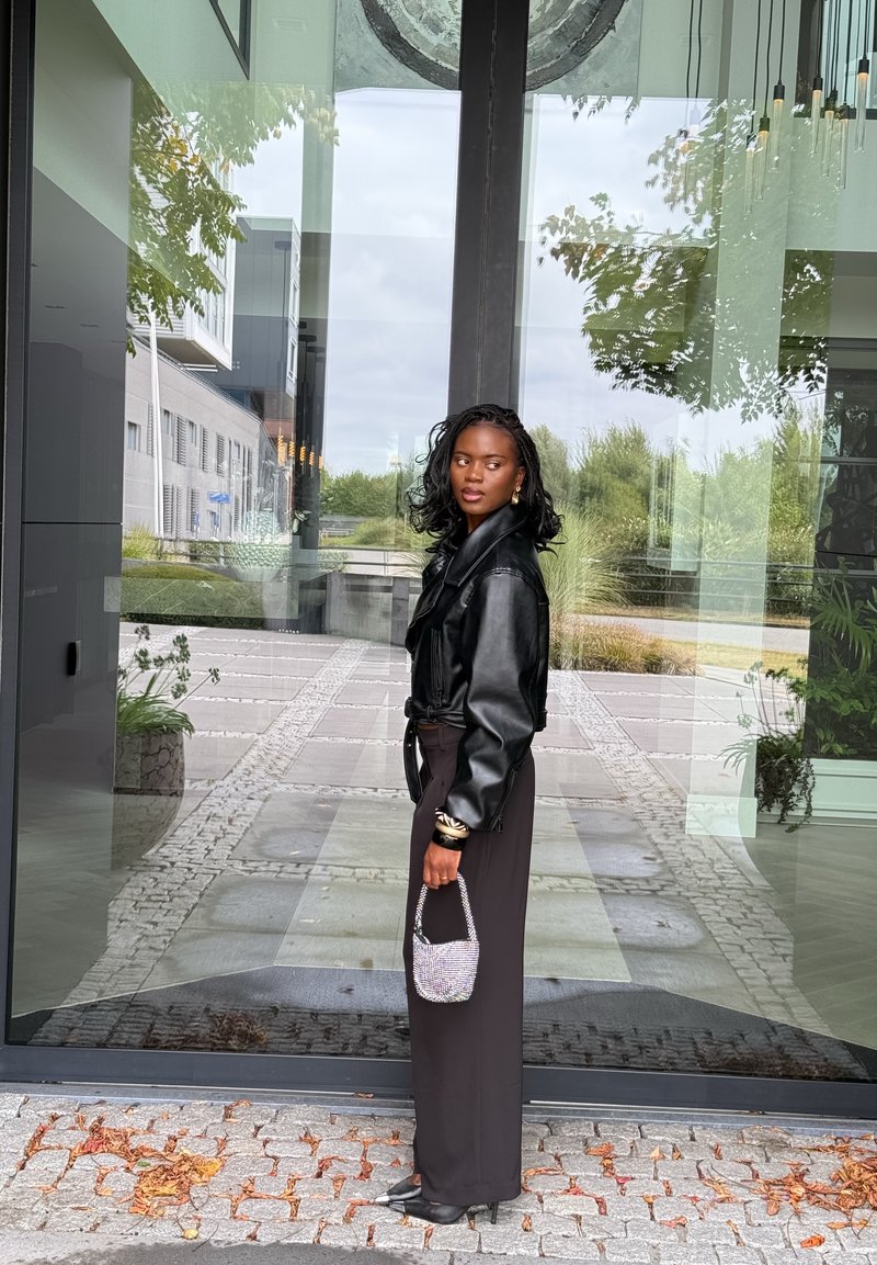 Svart läderjacka, långa svarta byxor, silverarmband som accent, håller en liten glittrig handväska. Glasdörr och stenlagd gångväg i bakgrunden.