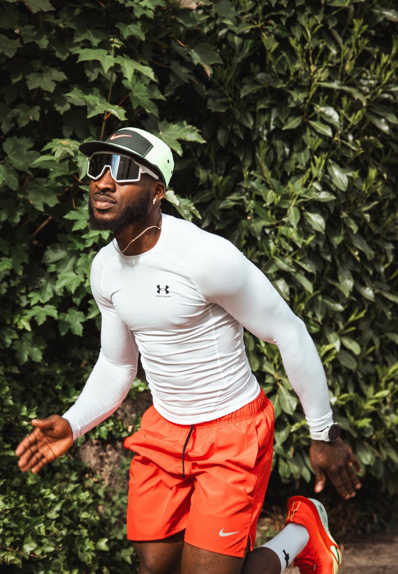 Homme athlétique portant un t-shirt blanc à manches longues, un short orange vif, une casquette néon et des lunettes de soleil en train de jogger à l'extérieur près de buissons verts feuillus.