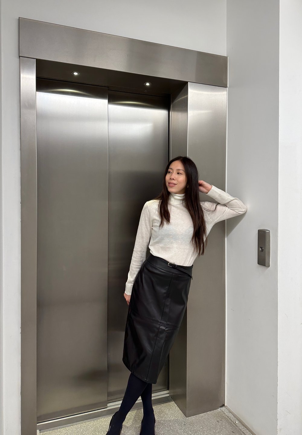Un lift din oțel inoxidabil cu două uși. O persoană stă lângă el purtând un pulover crem cu guler înalt și o fustă tip creion din piele neagră.