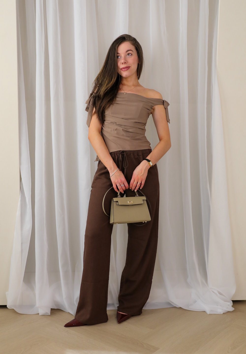 Off-Shoulder, hellbraunes Oberteil kombiniert mit dunkelbraunen, lockeren Hosen. Trägt eine kleine, olivgrüne Handtasche und elegante bordeauxrote Pumps.